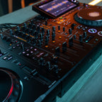 AlphaTheta DJM-V5 DJ mixer with professional controls, display and sleek black design, shown in a lifestyle setting.