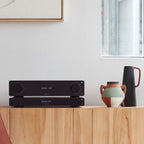 Arcam A15+ stereo amplifier in a stylish black finish, shown in a lifestyle setting with audio equipment and home decor.